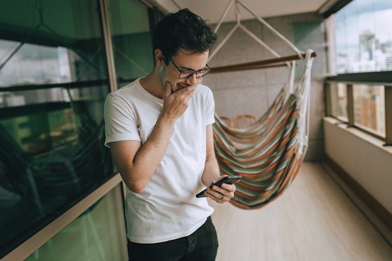 A man in casual attire checking his smartphone on a balcony with a hammock.